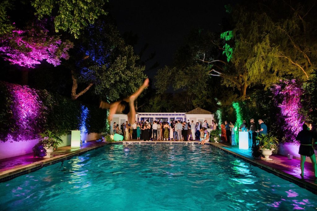 Backyard Wedding Inspo: an evening shot of a private residence's pool area with multicolored lights and people dancing on a dance floor